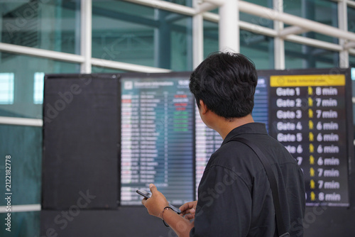 Asian man traveler looking and checking flight schedule departures board while holding a smartphone, travel concept, side view