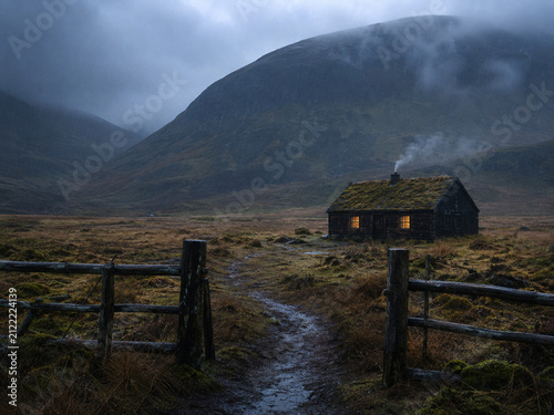 Old house in the mountains. Foggy mountain landscape with small cozy cottage with chimney. Ireland evening landscape. Warm light in windows. Farm in rainy hills. Hut with moss roof and old fence. 