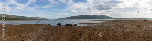 Wide panoramic landscape of a dried reservoir bed with dead wood and distant mountains