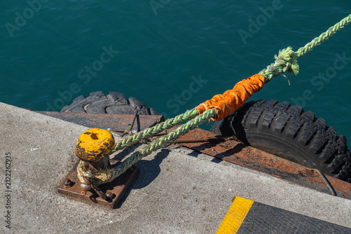 An old and rusty mooring bollard. A rope attached to the mooring bollard. Concrete pier.
