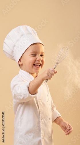 Happy little boy dressed as a professional chef holding a whisk with flour dust in the air, laughing and playing on a neutral studio background.
