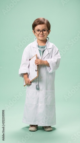 Cute little boy dressed as a doctor in an oversized white lab coat holding a clipboard with a drawing, standing against a green studio background, representing future career dreams and healthcare.