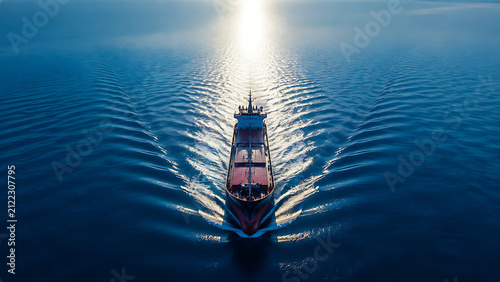 Aerial view of cargo ship crossing deep blue ocean with symmetrical wake trails and dramatic sunlight reflection on calm water
