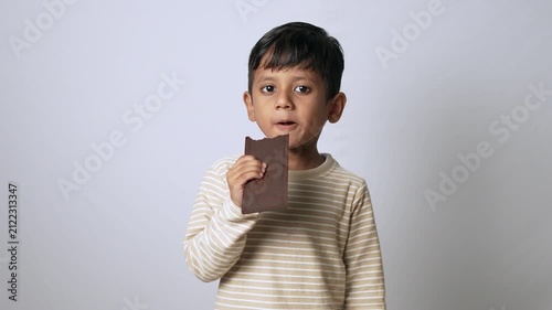 Indian boy eating chocolate bar. Happy child enjoying sweet snack on white background.