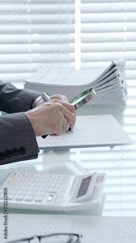 Hand holding magnifying glass and pen, studying financial paperwork and reviewing an invoice with a calculator on desk, performing a detailed audit and verification of data
