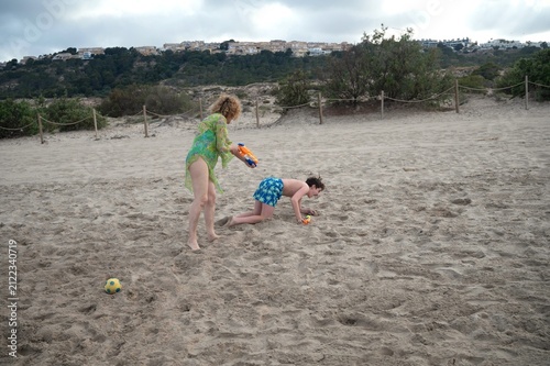 Mother playfully chasing her teenage son with water guns during a fun family day on a sandy beach