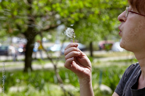 Profile of a woman blowing the seeds from a dandelion, symbolizing making a wish. Woman enjoying a sunny day outdoors, blows a dandelion at city park