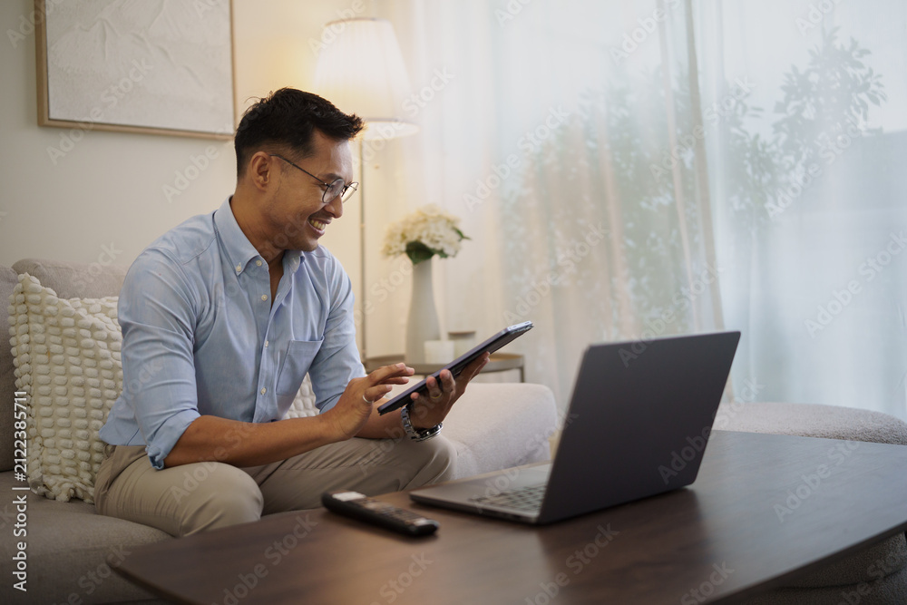 Fototapeta premium Smiling businessman using a digital tablet and laptop while relaxing on a sofa in a modern living room.