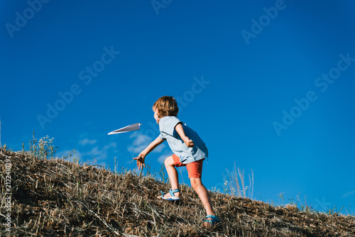 фотографія A happy young boy throws a white paper airplane into the air while standing on a grassy hill