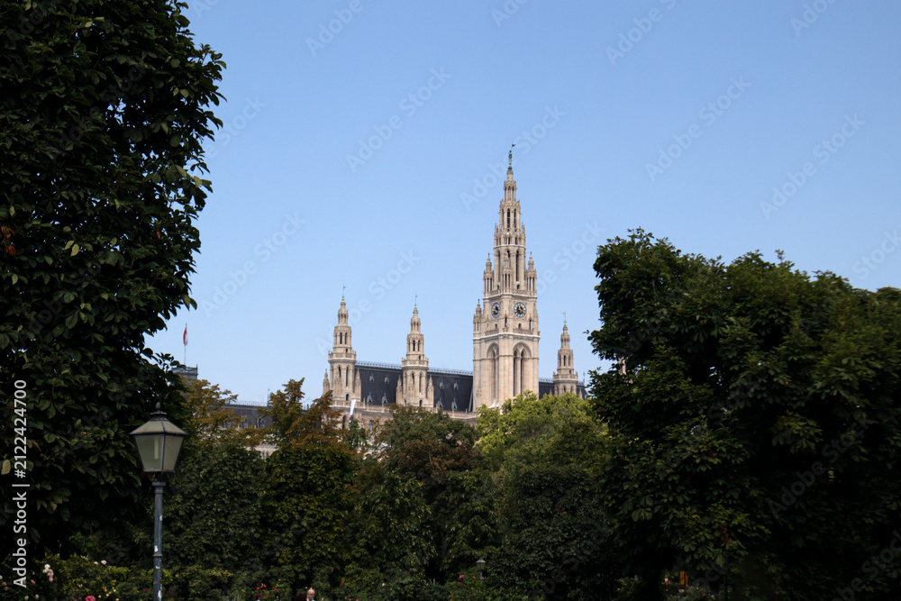 Obraz premium La maestosa torre neogotica del Municipio di Vienna Rathaus che svetta sopra gli alberi del parco Rathauspark con passanti lungo il sentiero in una giornata estiva, Austria.