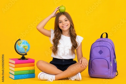 Smiling young girl sitting with globe and books and purple backpack on yellow background, back to school concept for educational advertising materials