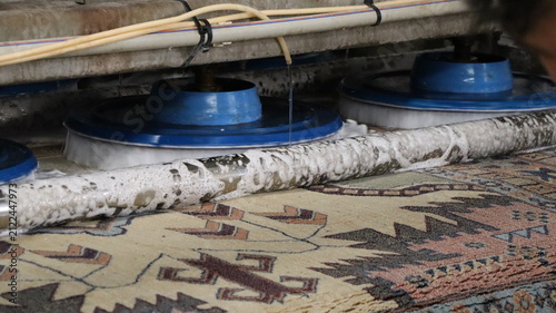 Close view of industrial carpet cleaning machine with rotating blue scrubbers working across patterned rug, showing foam, water flow, wet fibers, and low angle detail during commercial maintenance