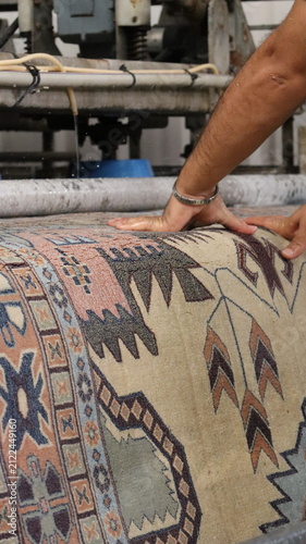 Worker using both hands to press large patterned rug onto cleaning machine roller in industrial workshop, close view revealing textured woven fibers, muted geometric motifs in blue, peach, and beige