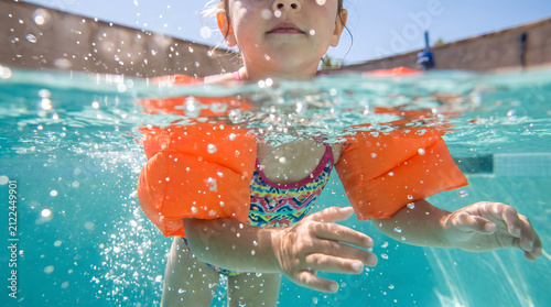 Little girl swimming underwater wearing orange floaties in a pool with bubbles and sunlight