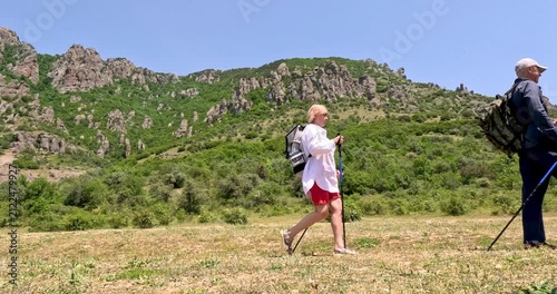 Man and a woman walk with walking poles and backpacks on the grass against a backdrop of mountain peaks.