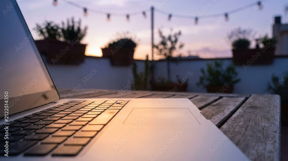 Fototapeta premium Laptop rooftop sunset table keyboard touchpad terrace evening string light wood blurred background outdoor twilight open screen device workspace