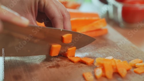 Woman hand dicing carrot with kitchen knife on wooden cutting board.