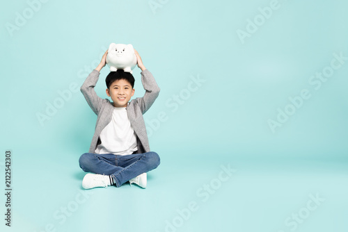A cheerful Asian boy sitting cross-legged with a white piggy bank on his head in studio, illustrating money saving, childhood financial planning, and education fund concept with copy space