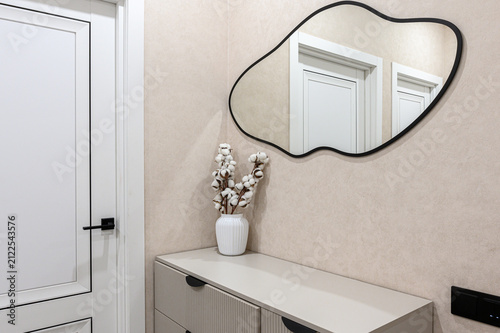 modern entryway: a beige console with vertical grooves, a white vase with cotton branches, an irregular-shaped black-framed mirror, and a white door with a black handle