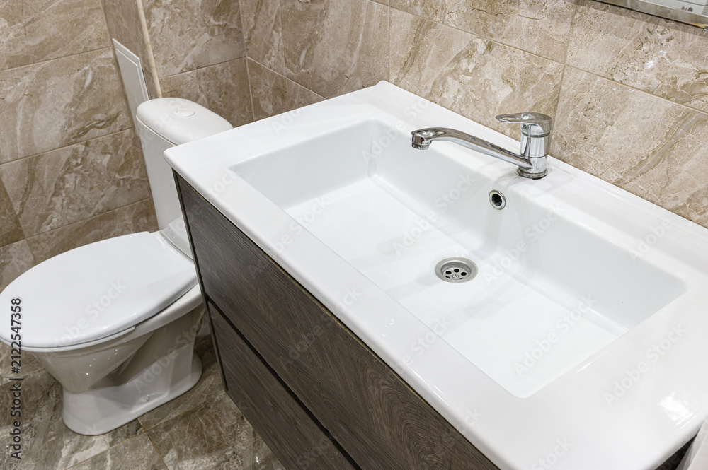 Fototapeta premium compact bathroom with a white rectangular sink atop a grey wood-grain vanity. chrome faucet, white toilet, and beige marble-patterned tiles complete the setup