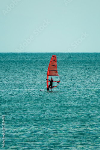 Obraz na plátne A windsurfer glides across the calm sea with an orange sail under an overcast sky
