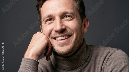 Smiling handsome middle-aged man in a brown turtleneck looking away with a genuine cheerful expression. Studio close-up portrait on a dark background conveying optimism, confidence, positive emotions