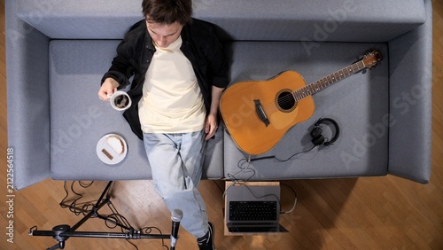 Canvas Print Young man relaxing on a couch, holding a coffee cup next to an acoustic guitar,