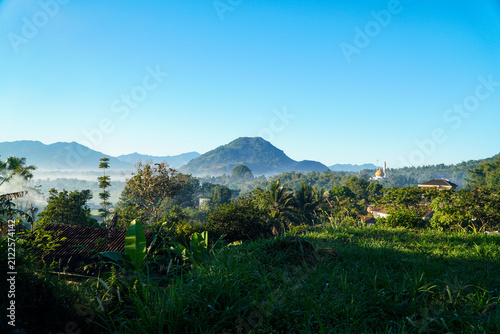 Bild auf Leinwand Vibrant green hills and misty valleys under clear blue sky with distant mountains and a golden-domed mosque nestled among trees