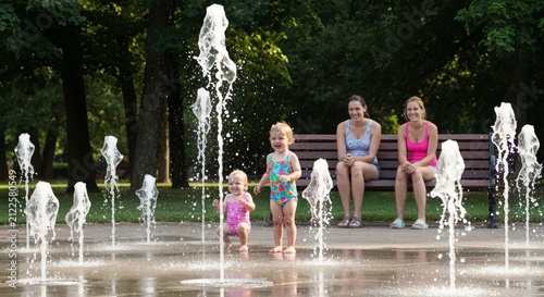 Toddlers squealing at gentle fountains, parents watching nearby, rubber sandals and bright swimsuits