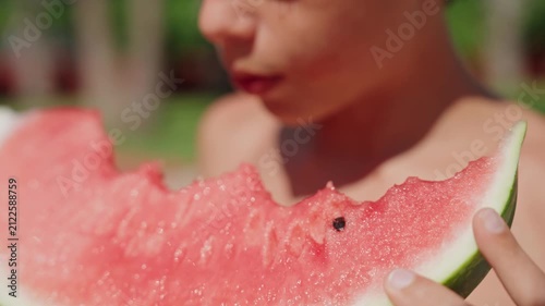 A close-up of a young boy taking a bite of a large, juicy slice of fresh watermelon on a sunny summer day. The vibrant red fruit, visible texture, and refreshing juice highlight healthy seasonal