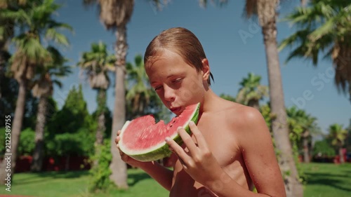 A happy young boy enjoys a large slice of fresh watermelon outdoors on a sunny summer day surrounded by palm trees and tropical greenery. Standing shirtless at a resort, he is about to take a bite of