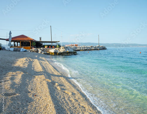 Bild auf Leinwand A quiet pebble beach with crystal clear turquoise sea water, featuring a small red-roofed building and a stone pier under a clear blue sky during summer morning