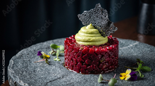 A beautifully detailed photorealistic stock photograph capturing an avant-garde beetroot tartare with avocado mousse and a charcoal crisp on a textured grey stone plate.