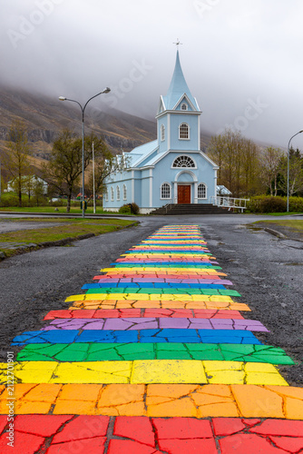 Blue Church Seydisfjardarkirkja behind a rainbow painted pavement street in Seydisfjordur, Iceland