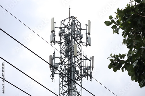 A metal lattice telecommunications and cellular tower against an overcast sky, with power lines and green foliage framing the sides.