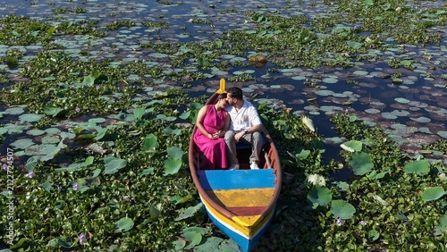Quadro su tela Aerial drone view of a couple enjoying a colorful boat ride across a scenic lake covered with lotus leaves and aquatic plants, surrounded by lush greenery