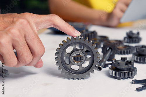 Close-up of an engineer's hand holding a metal cogwheel over a desk scattered with mechanical parts. Concept of industrial teamwork, mechanical engineering, innovation, and product design.