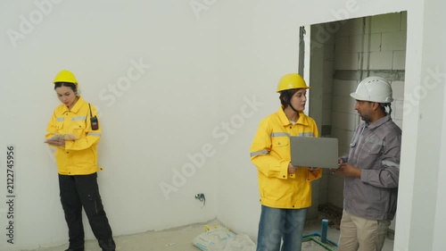Home inspector team in unfinished building room with hard hat and clipboard, reviewing wall plan, worker discussion, site inspection for construction project
