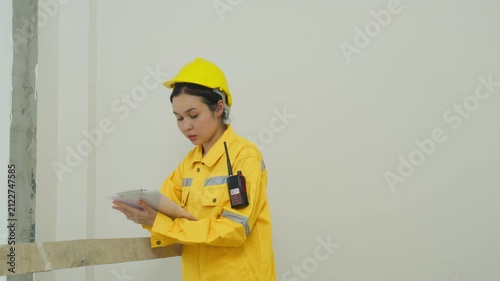 Home inspector examining wall surface in bright interior, wearing yellow helmet and jacket, checking clipboard inside empty room, focused professional worker in building interior