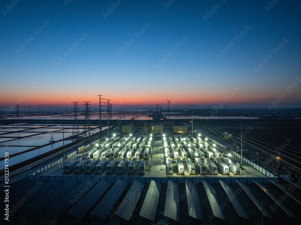 Fototapeta premium Energy storage station and photovoltaic array under twilight sky with illuminated equipment and coastal wetlands