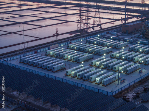 Naklejka Large-scale energy storage station and photovoltaic array at dusk with illuminated modules and surrounding industrial infrastructure