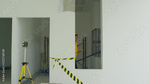 Interior construction site survey with worker safety helmet, caution tape, tripod inspection, unfinished wall renovation and checklist review for indoor safety site assessment