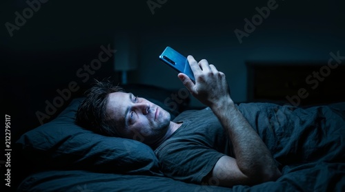 A man lies awake in bed late at night, his face lit by the harsh blue light of his phone screen, illustrating digital burnout, insomnia, and the compulsive habit of late-night scrolling
