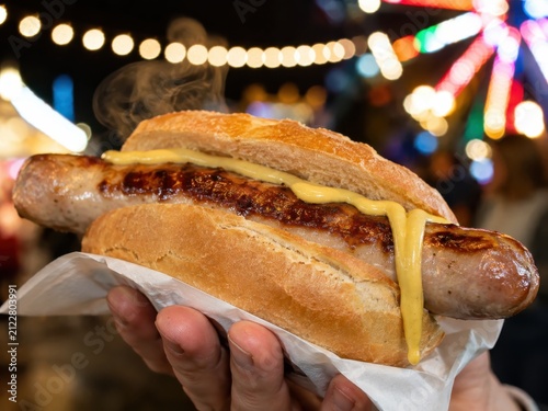 Gegrillte Bratwurst im Brötchen mit tropfendem Senf – Dampf, Grillspuren und bunte Volksfestlichter machen den deutschen Imbiss besonders appetitlich und traditionsreich.