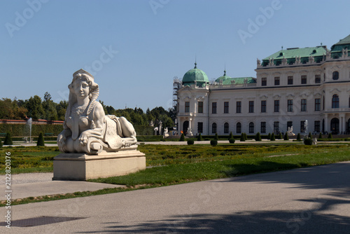 Statua monumentale di una sfinge in pietra nei giardini barocchi con la facciata del Palazzo del Belvedere Superiore sullo sfondo, famosa attrazione storica a Vienna, Austria.