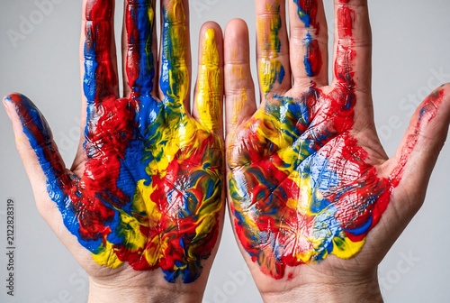 Close-up of two open palms coated with thick red, blue and yellow acrylic paint. Vivid, messy texture on skin over a neutral gray backdrop, perfect for creativity, education, art, and DIY concepts.