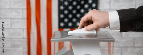 Voter casting a voting ballot into a transparent ballot box with the US flag. American elections, democracy and voting rights.