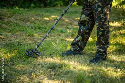 Photography A person stands on grass in a wooded area, using a metal detector to search for buried items