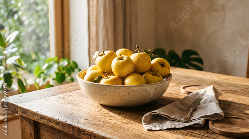 Apples rest in a ceramic bowl on a weathered wooden table near a sunlit window with a linen napkin placed beside the fruit in a cozy kitchen setting that emphasizes natural light and home comfort.