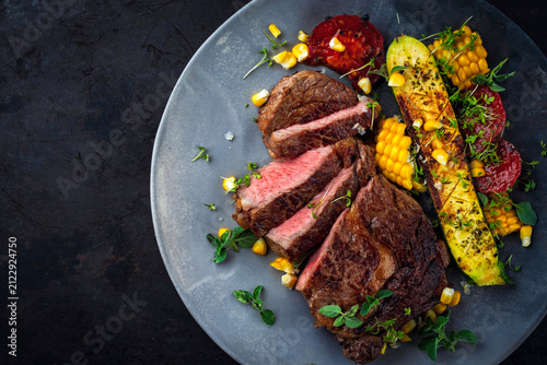Dry aged roasted beef entrecote with grilled vegetables, zucchini, sweet corn and greens served as top view on a design plate with copy space right